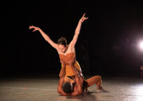 A dancer laying on the stage holds up another by the ribs who has her arms stretched in a V.