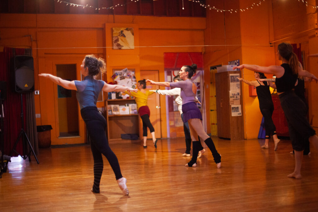 Bianca demonstrates a tendu derriere to beginning level adults behind her in a ballet studio.