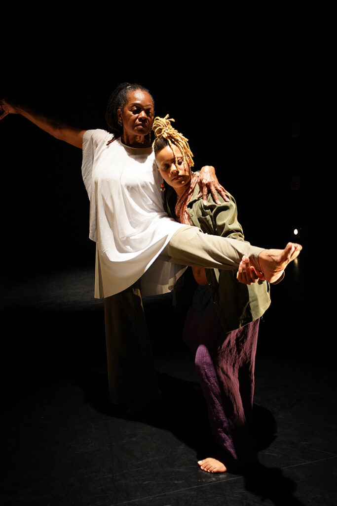 One dancer leans against another dancer who is lifting their leg. The background is black.