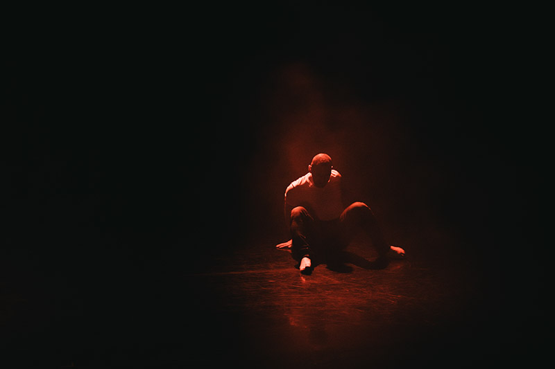 Stuart sits with his legs bent in front of him. He is on a dimly lit stage and all that is visible is a reddish silhouette.