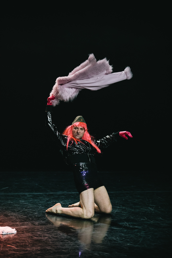 Stuart is kneeling onstage in a leather jumper and pink hair, swirling a frilly jacket above his head.