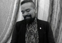 A black and white headshot of Jose smiling and looking down. He is wearing a patterend shirt and a jacket.