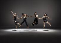Four mixed-race Asian American dancers stretched out horizontally holding hands, jumping up in unison under a pool of light.