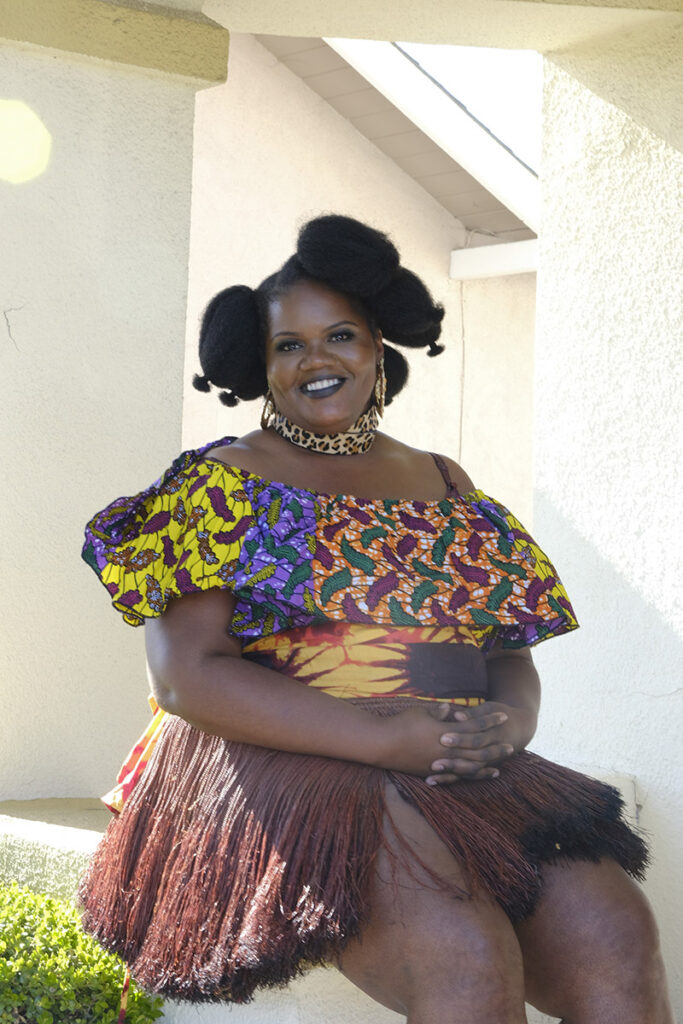 A headshot of Muisi-kongo sitting outside and wearing a multi-colored shirt and grass skirt.