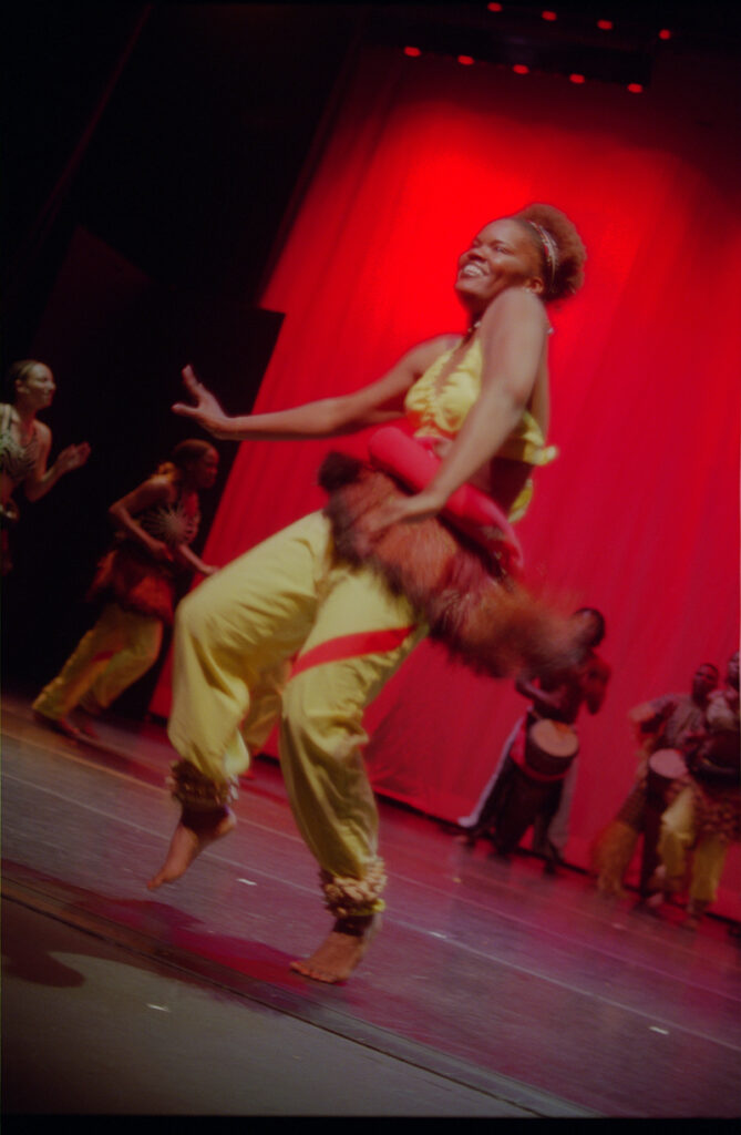 Muisi-kongo and others dance on a red-lit stage wearing yellow and red costumes.
