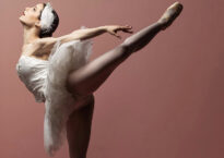 Sydney in a dramatic arabesque wearing a white swan tutu against a pink backdrop.