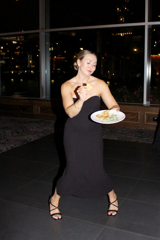 Alexandra wearing a black dress and holding a plate at a party. She jokingly makes a funny pose holding a chip.