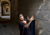 Shebana is in a stone tunnel. Light falls on her from a shaft and she artfully crosses her arms in front of her,