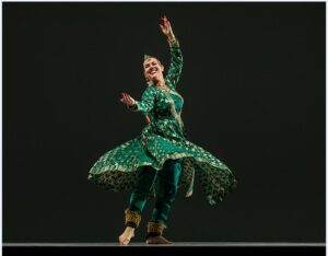 Charlotte stepping back and raising her arms wearing a green and gold dress