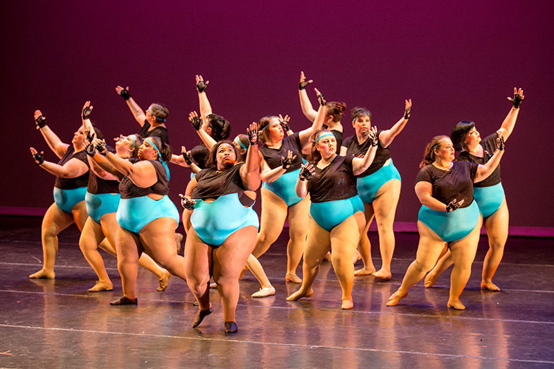 13 dancers onstage gracefully reach upward, all wearing blue leotards and black t-shirts