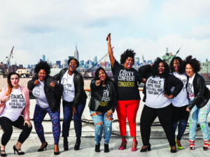 Eight fat women in a row smile and cheer with a city skyline in the background.