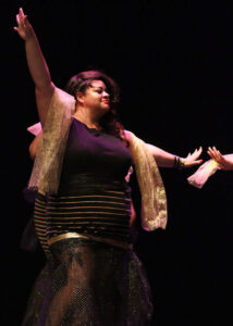 Tigress wearing a black and gold dress with one arm up and one arm to the side.