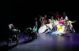 Two wheelchair dancers are in the foreground on stage and a clump of colorfully dancers make a shape together in the background.