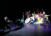 Two wheelchair dancers are in the foreground on stage and a clump of colorfully dancers make a shape together in the background.
