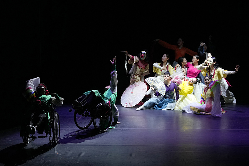 Two wheelchair dancers are in the foreground on stage and a clump of colorfully dressed dancers make a shape together in the background.