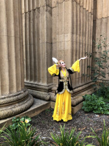 Natalie posed with both arms around her head standing by a large column outside and wearing a long yellow dress.