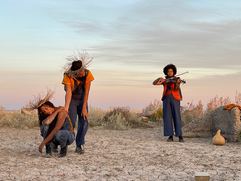 Nikesha squats down to the left of the frame, Miles stands behind her, and Lazarus plays the violin to the right of the frame. They are in a desolate desert.
