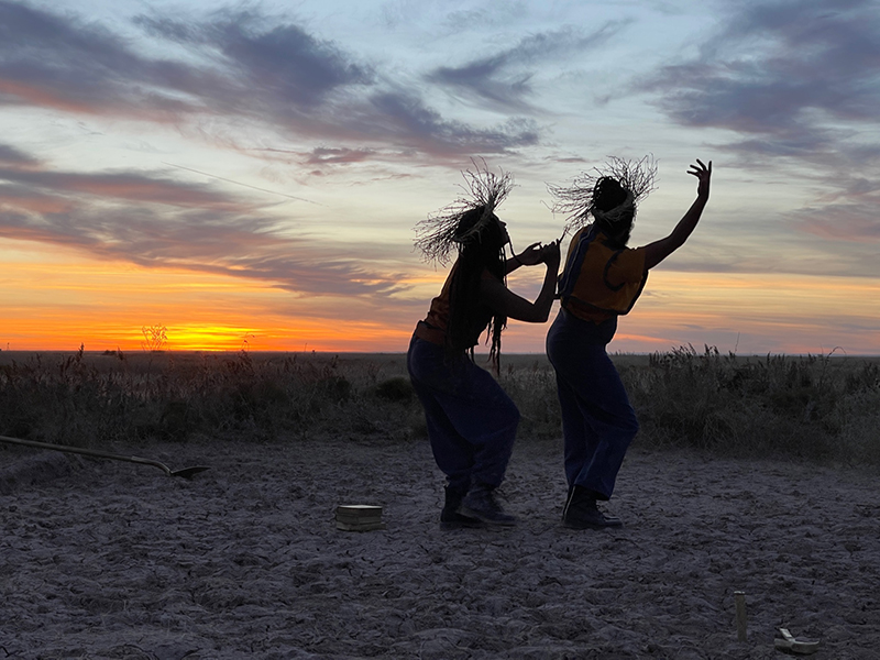 Two dancers dance in silhouette against a sunset in the desert wearing straw hats.