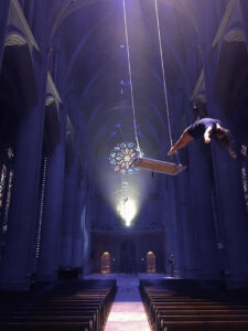 One dancer leans into a backbend off an enormous swing while another dancer in the background climbs a very tall ladder at Grace Cathdral.
