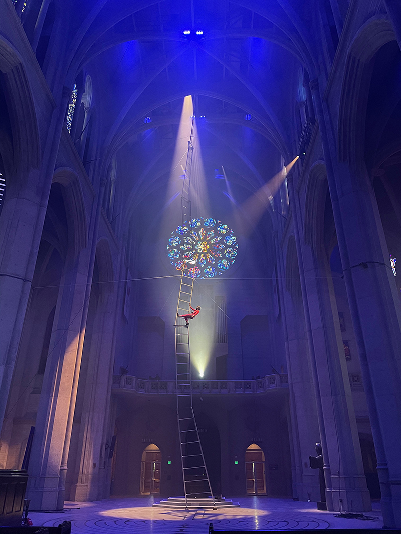 A dancer is bathed in stage light while climbing an enormous ladder that extends to the ceiling of Grace Cathdral.