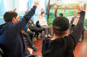 People sitting in chairs in a circle with one arm raised.