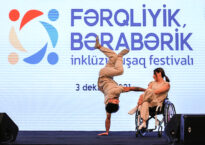 One dancer in a wheelchair and one dancer doing a handstand are in front of an Azerbaijani sign.