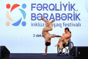 One dancer in a wheelchair and one dancer doing a handstand are in front of an Azerbaijani sign.