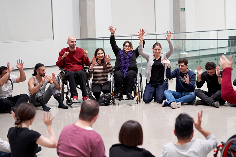 Dancers with and without disabilities sit in a circle and raise both hands.