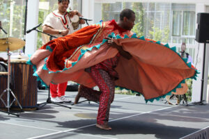 Etienne swirls around in a large round orange and red costume. A drummer plays behind him.