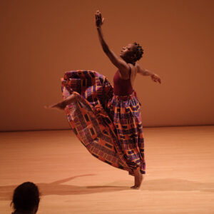 Rujeko on a wooden floor performing with one arm raised and one leg raised. She is wearing a long skirt.