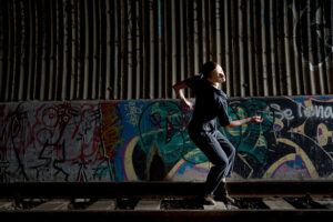 Emily is pictured to the side with knees and arms bent. A mural is dimly lit behind her.