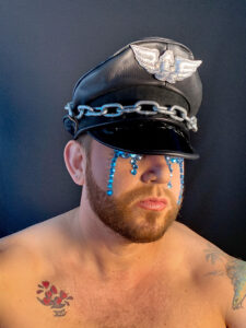 Kevin close-up topless wearing a military style hat over their eyes and blue glitter droplets down their cheeks.