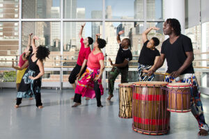 Students life one arm. A drummer is in the corner. High rise buildings are out the window behind them.