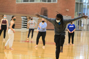 Momar is dancing with arms to the sides and one leg lifted. Students are watching in the background.