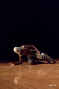 Momar is crouched low to the ground with one arm extended. He is shirtless with his shirt pulled over his face.