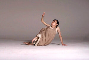 Photo of Kazuyo seated with elevated hand.