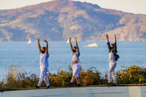 Three dancers walk in a line with their hands raised above their heads.
