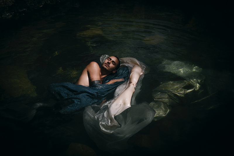 Illan floating in a dark pool with fabric floating with him.
