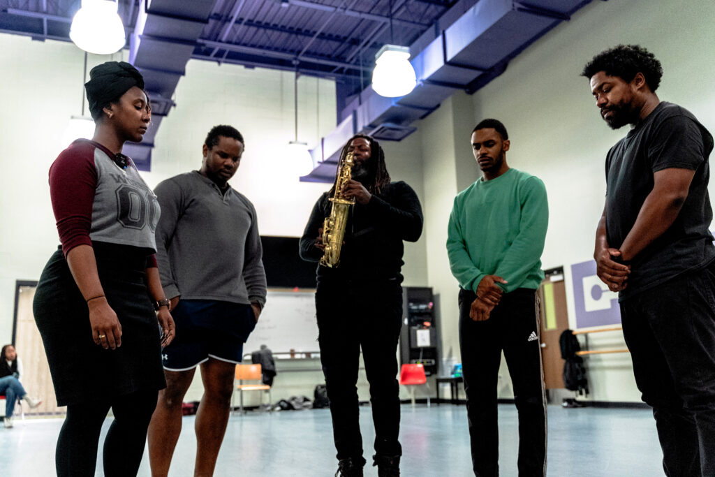 Five performers, one with a saxophone, stand in a circle.