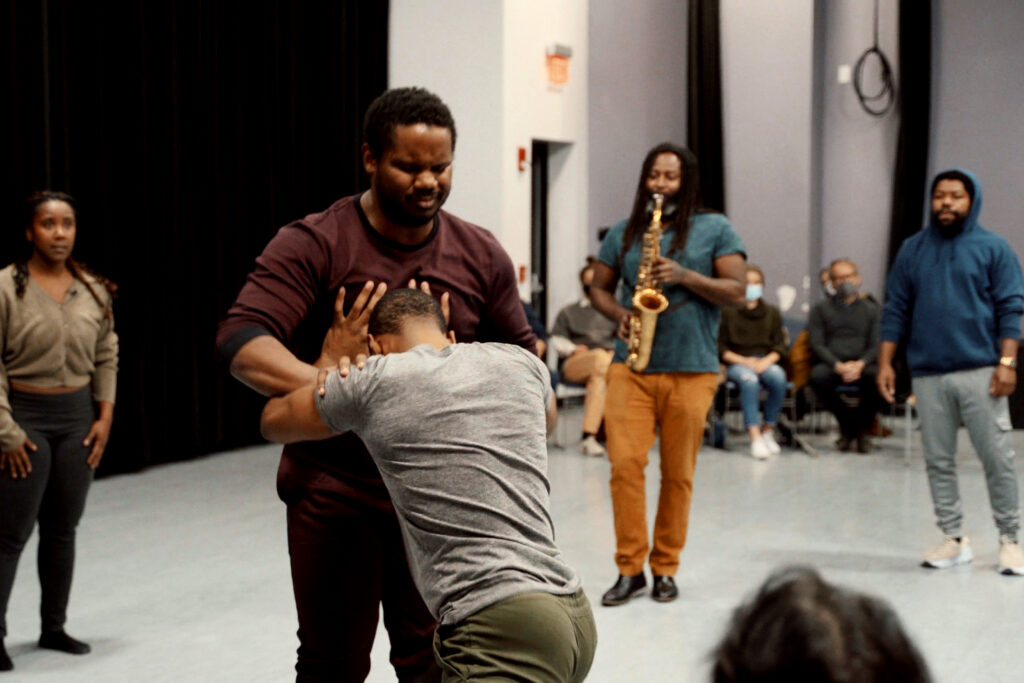 Two dancers wrestle while three performers in the background look on, one playing a saxophone.