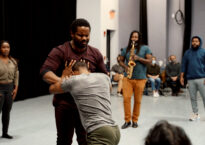 Two dancers wrestle while three performers in the background look on, one playing a saxophone.