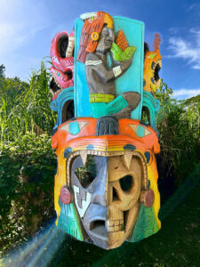 A wooden mask with two half faces and a figuring holding corn on top. A lush field is in the background.