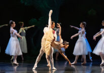 Michelle in a deep penchee with a partner. She is wearing a yellow tutu and dancers in white tutus move behind her.