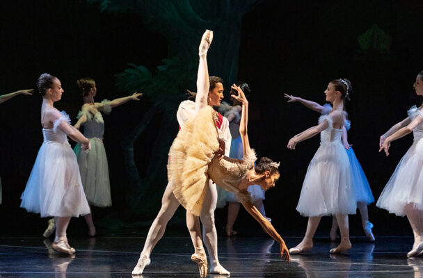 Michelle in a deep penchee with a partner. She is wearing a yellow tutu and dancers in white tutus move behind her.