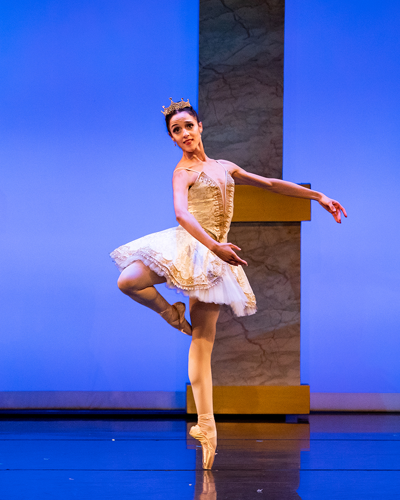 Michelle in passe on pointe wearing a gold tutu against blue stage lighting