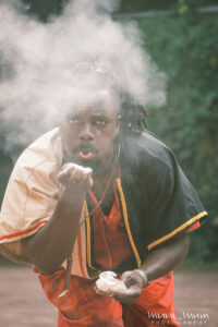 West blows chalk or dust at the camera, leaving him clouded behind the dust. He is wearing orange and black and is a little hunched over. Trees are in the background.