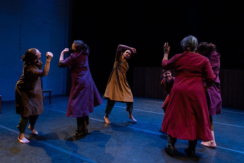 Dancers of different ages spread out on stage facing different directions and wearing different colored dresses.