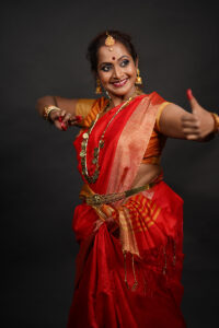 Yashoda wears a red sari with gold jewelry. One arm is extended in front of her with her thumb lifted. The other arm is near her chest bent at the embow.