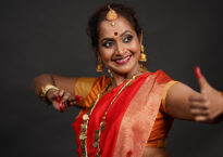Yashoda wears a red sari with gold jewelry. One arm is extended in front of her with her thumb lifted. The other arm is near her chest bent at the embow with two fingers extended.