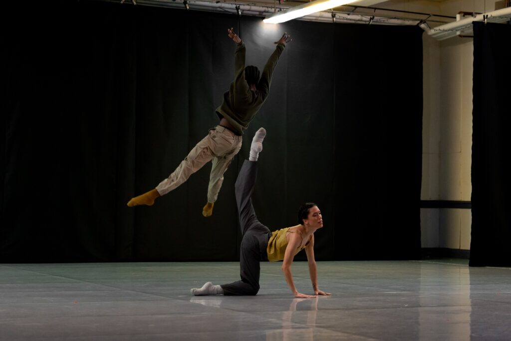 In a dance stuio, one dancer jumps high above another dancer who is on her hands and knees with her leg extended up behind her.